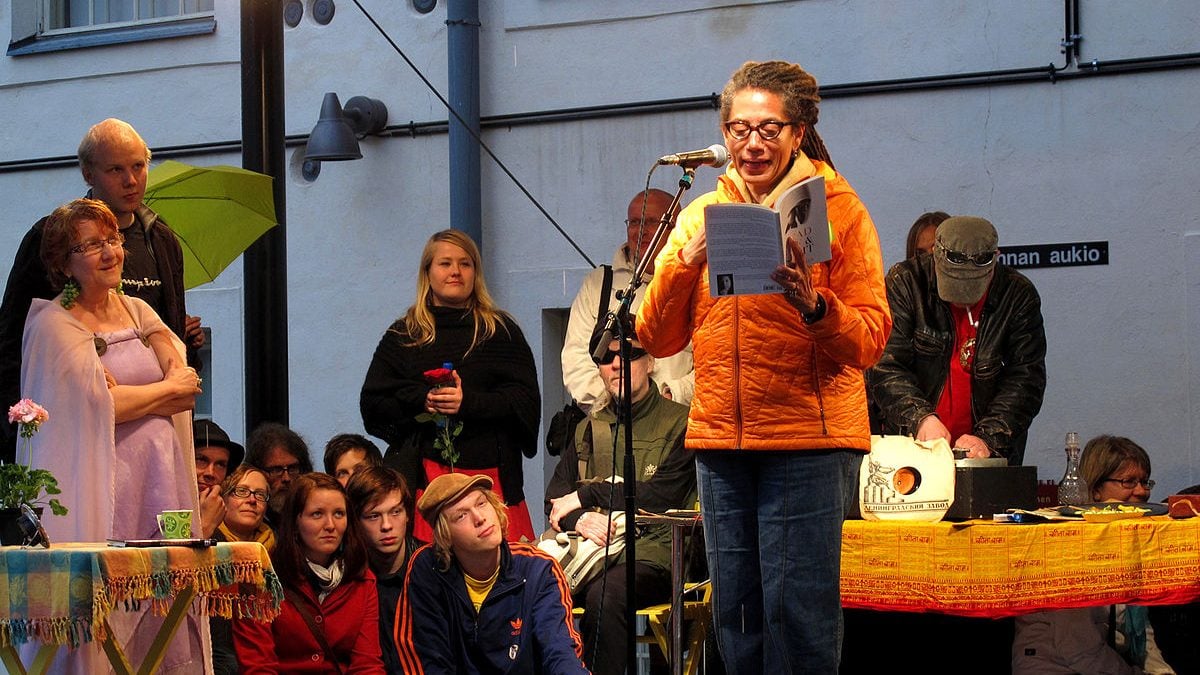 A woman reads from a book in front of a microphone to an audience.