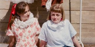 Two girls sit in front of a clothes rail, with white ruffles round their necks, one of them looking at the other