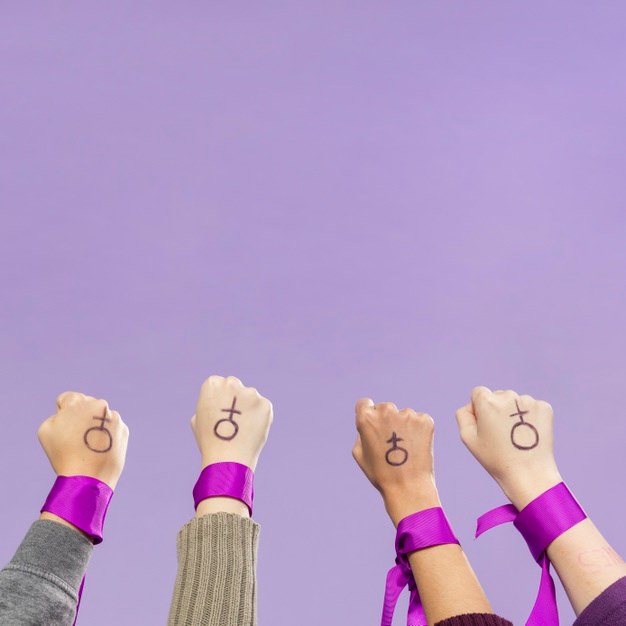 4 people hold up their fists against a purple background. They have pink ribbons tied around their wrists and the female sex symbol drawn on their hands.