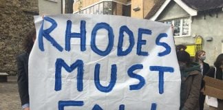 A white banner being held up by two people who one are hidden behind it. In blue letters is written: "RHODES MUST FALL"