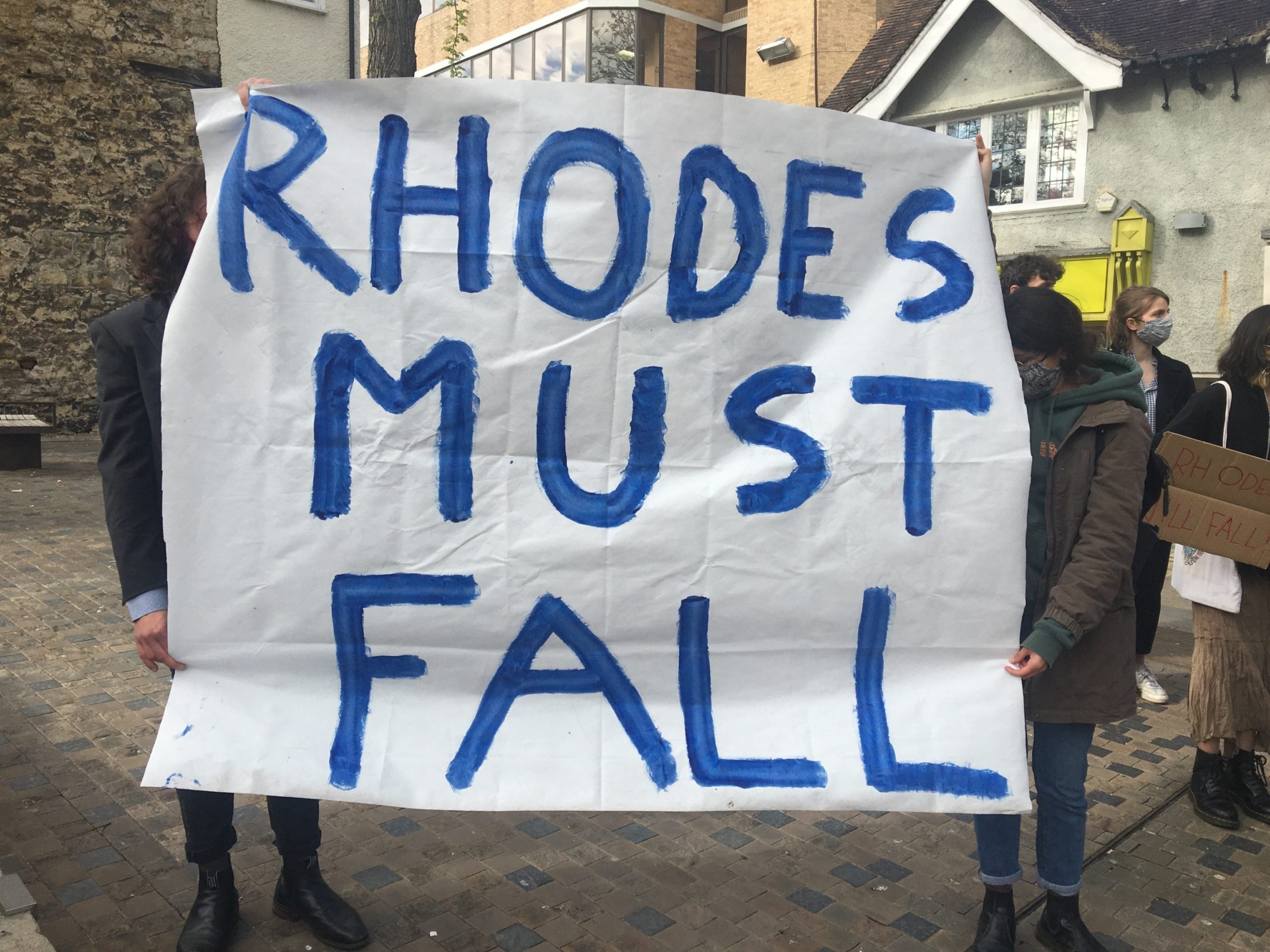 A white banner being held up by two people who one are hidden behind it. In blue letters is written: 