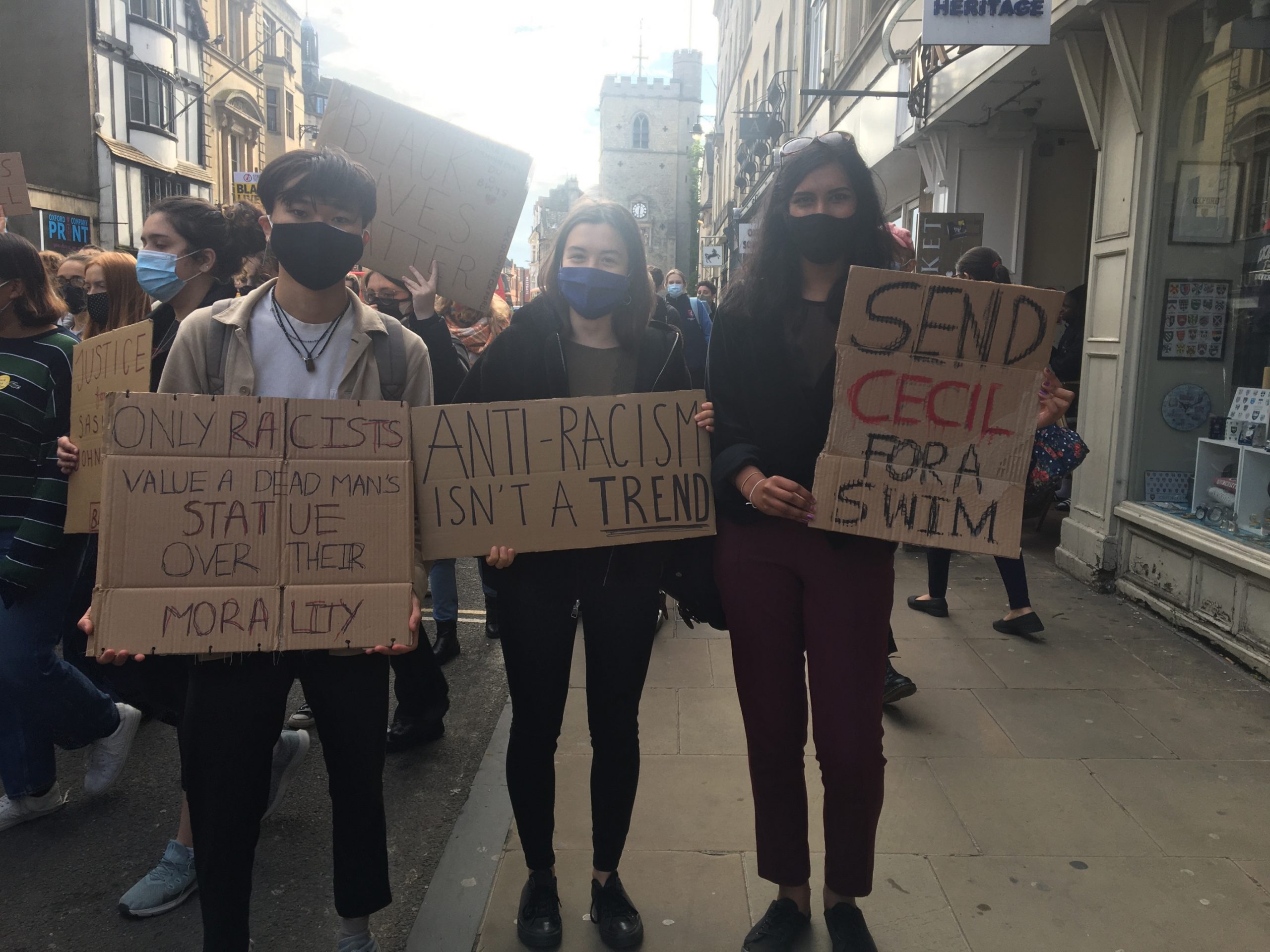 Three young adults wearing masks at a protest holding signs which say: "ONLY RACISTS WOULD VALUE A DEAD MAN'S STATUE OVER THEIR MORALITY", "ANTI-RACISM ISN'T A TREND" and "SEND CECIL FOR A SWIM".