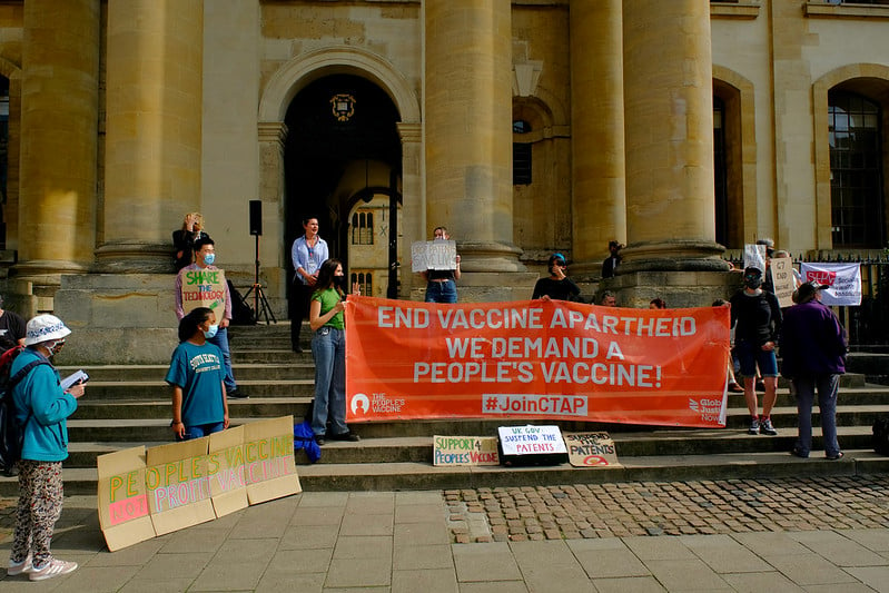 ID: Protestors standing on steps of an old building with beige/brown pillars. They hold a sign saying: END VACCINE APARTHEID / WE DEMAND A / PEOPLE'S VACCINE !