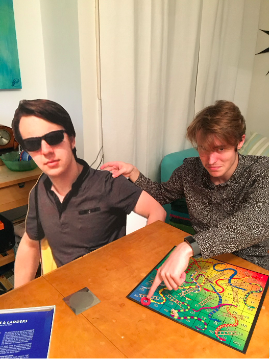 Two friends play snakes and ladders; one is a cardboard cut-out.