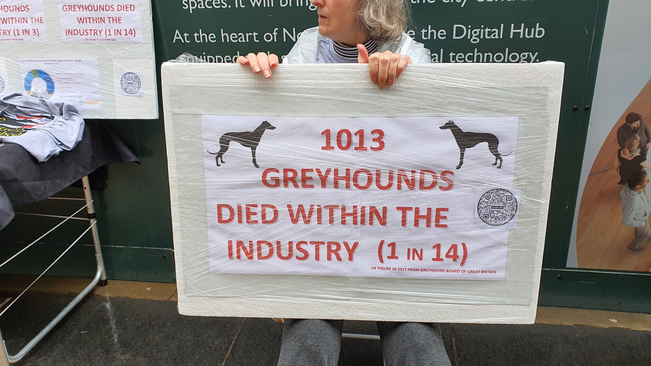 A women holding a sign saying: "1013 GREYHOUNDS DIED WITHIN THE INDUSTRY (1 in 14)" and in small writing at the bottom "UK FIGURE IN 2017 FROM GREYHOUND BOARD OF GREAT BRITAIN"