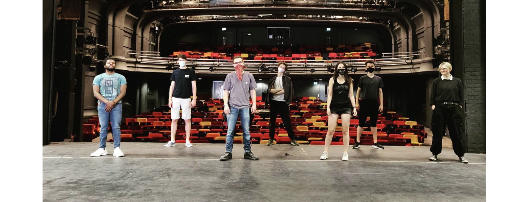 Seven people standing on a stage, facing away from an empty audience, whilst wearing masks.