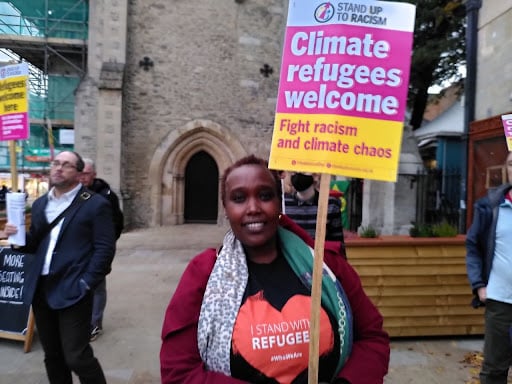 Caritas, from the Organisation Asylum Welcome holds a sign sying “Climate refugees welcome” and wears an organise T-shirt saying “I STAND WITH REFUGEES”. She stands in front of Carfax tower and there are three other protestors in the background.