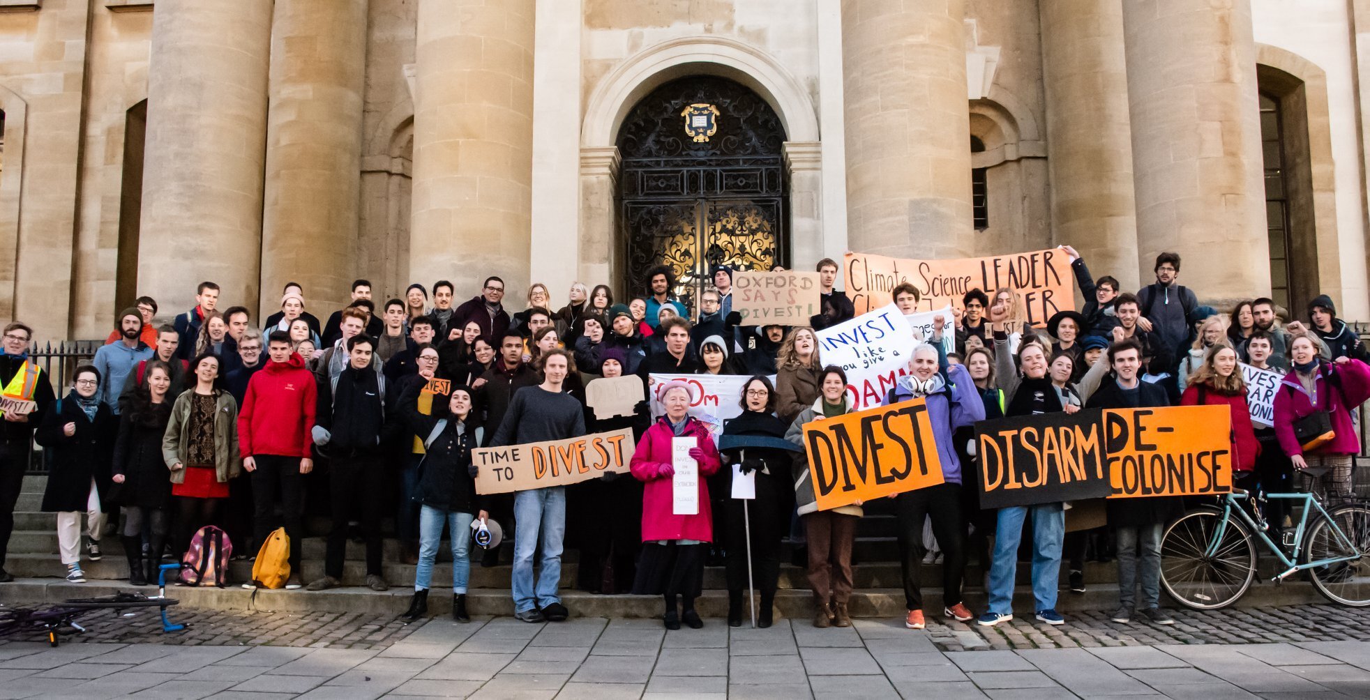 Many people outside an Oxford building holding signs calling for divestment, disarming and decolonisation.