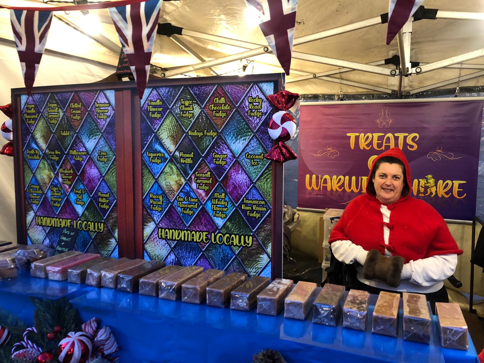 Liz sits at Treats of Warwickshire fudge stand, showcasing many bars of fudge and wearing red hooded top.