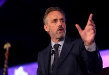 Jordan Peterson stands at a lectern, his left hand reaching out.