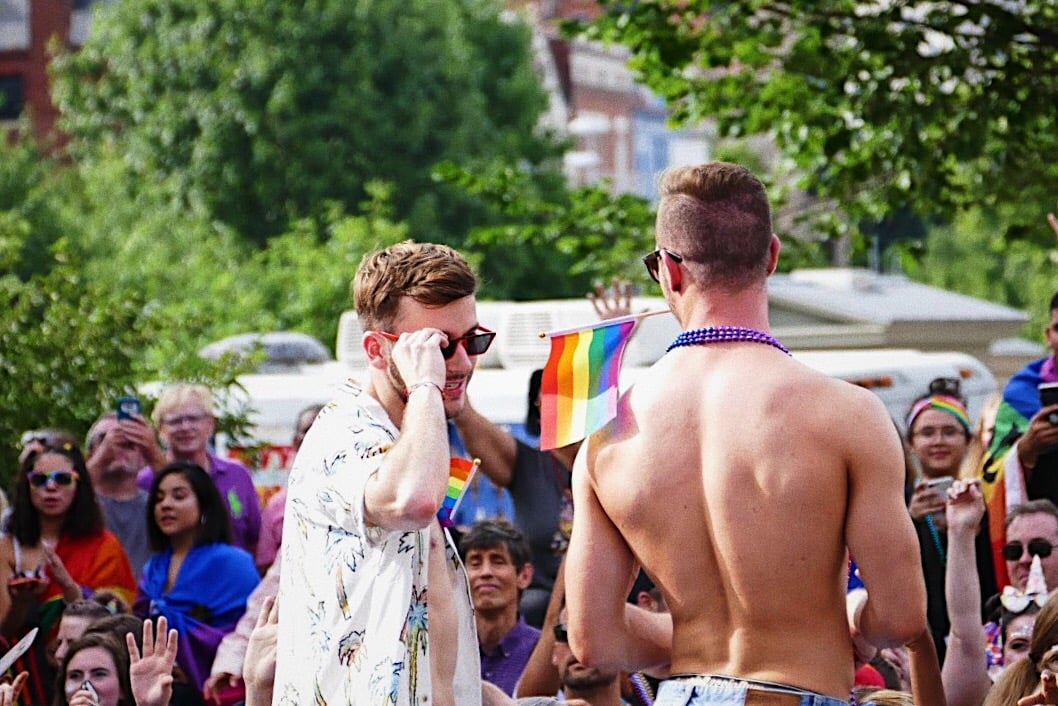 Image of two men at a Pride parade, one of whom is holding the LGBTQ+ pride flag.