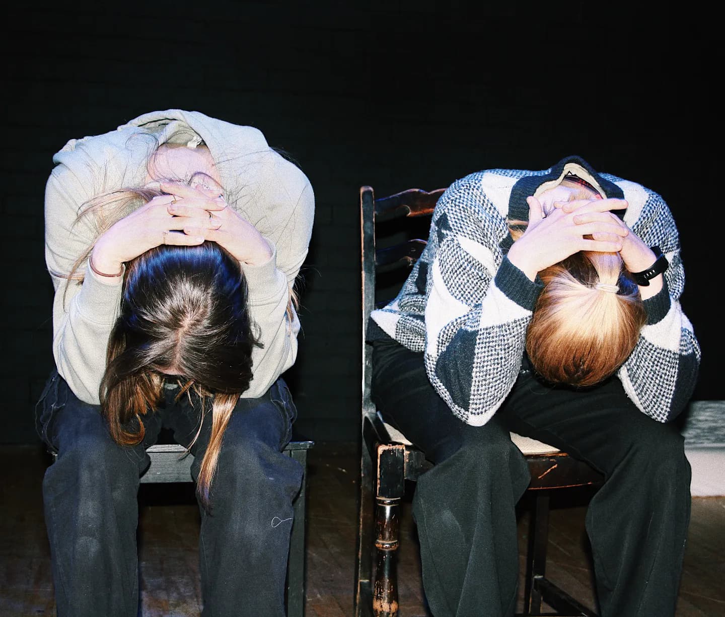 Two women sit side by side on chairs with their heads down and their hands on the backs of their necks