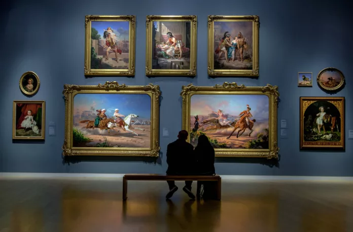 alt= a couple on a date sitting in front of a wall of paintings in an art gallery