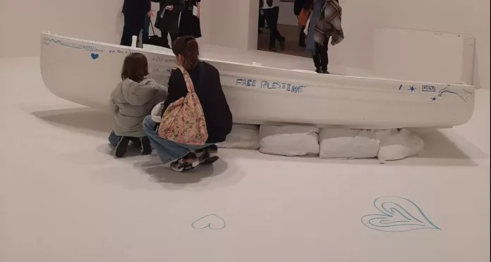 A mother and a child crouched in front of a white boat in a white room. The boat has blue writing on it. The girl is writing on the boat.