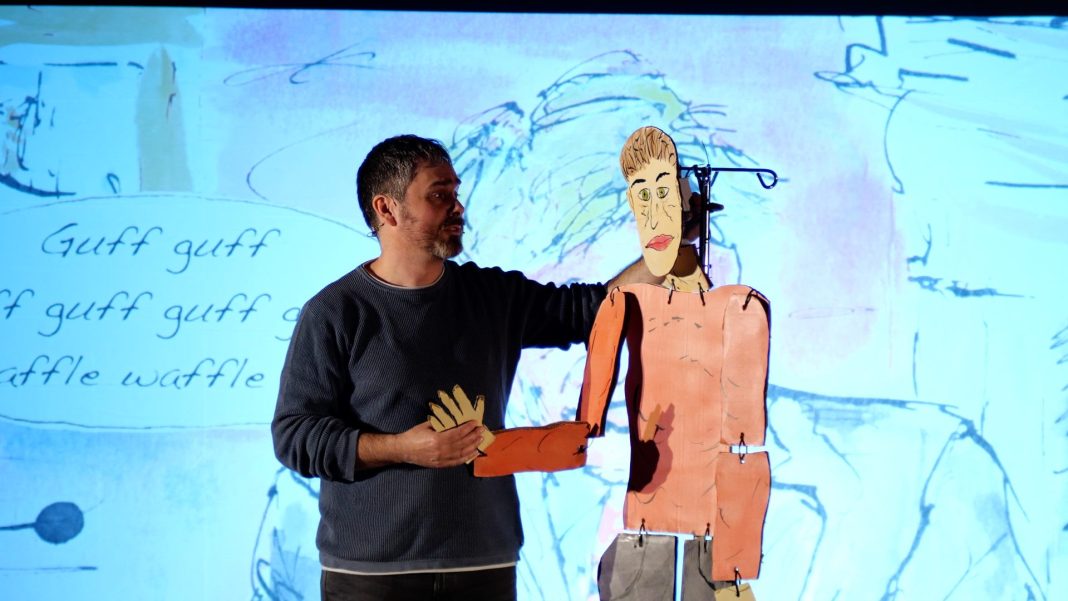 John Butler holding man puppet in front of illustrated backdrop.